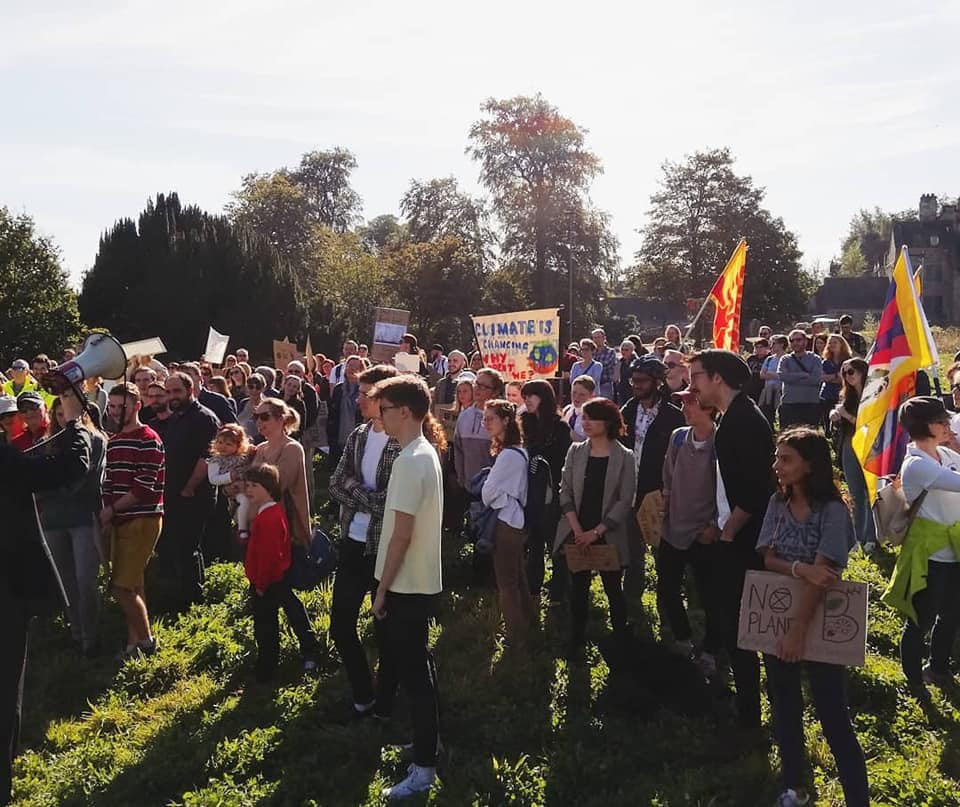 A group of 100+ strikers of all ages standing in a field in the sun, listening to a speech.