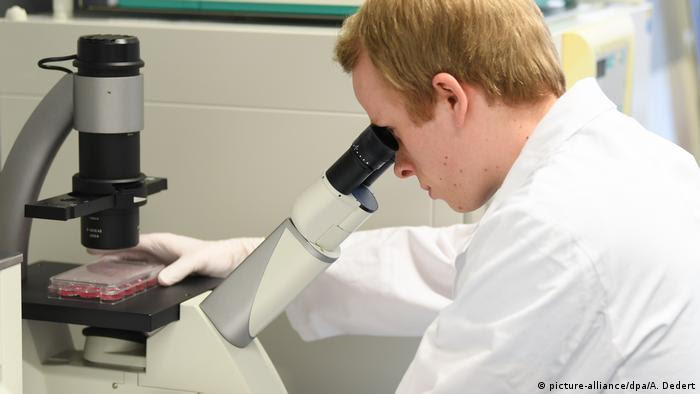 Homem de jaleco branco e luvas se debruça sobre microscópio com amostras em laboratório