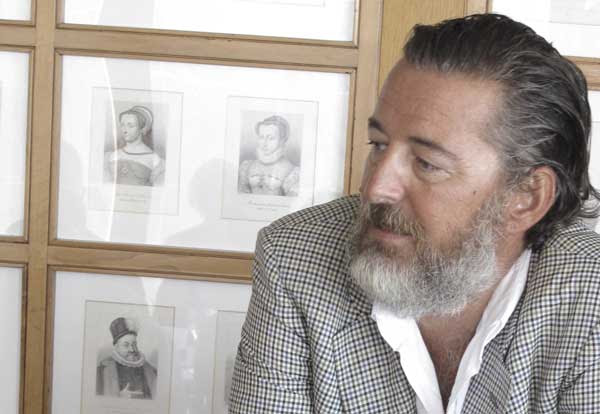 Photo of Sebastiano Rosa - producer of Barrua Isola dei Nuraghi IGT, 2016, showing his head and shoulders, informally dressed in an open white shirt and sports jacket looking to the left in front of a series of small lithographs of two women and one man.