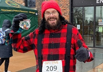 A runner decked out in red buffalo check flannel and suspenders grins at the camera while holding a finisher's medal triumphantly.