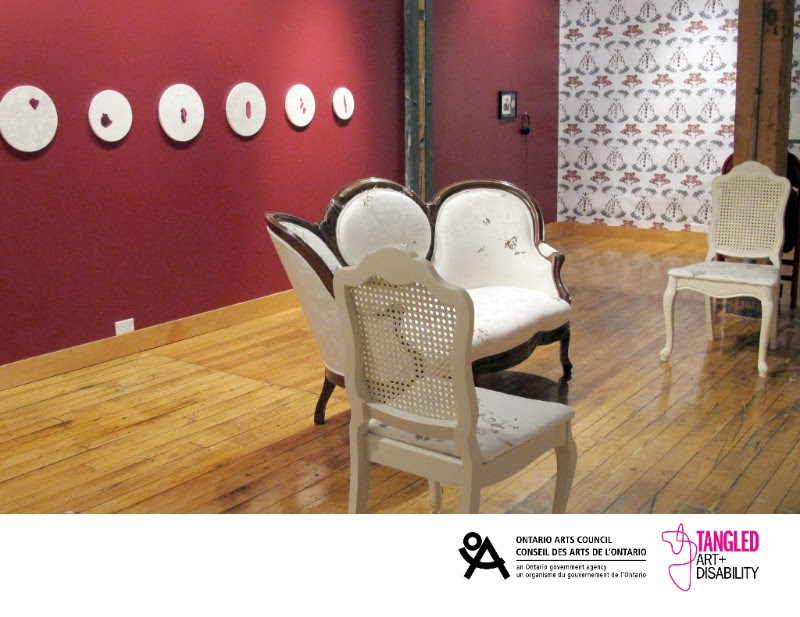 A photograph of Tangled Art Gallery with Vanessa Dion Fletcher’s exhibition, Own Your Cervix. The gallery features one red wall one wall with printed wall paper, and wooden floors. Hanging on the red wall are six white circles with organic red shapes adorned on them. On the gallery floor is a set of 3 chairs, two white wicker backed chairs and one victorian era couch with white printed upholstery and a dark wooden frame. Below the image is the logo for the Ontario Arts Council and Tangled Art + Disability Logos. 