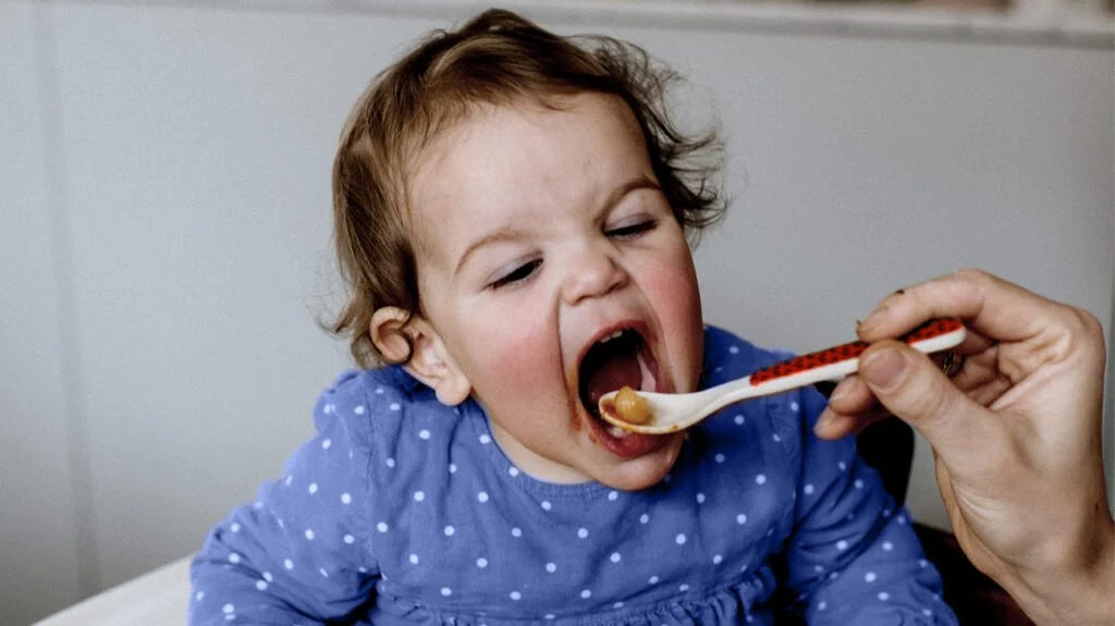 A toddler with their mouth open being fed food on a spoon