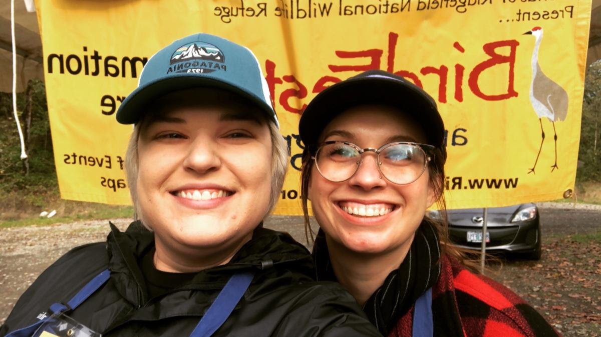 Two board members volunteering for BirdFest & Bluegrass take a selfie in front of a BirdFest & Bluegrass banner