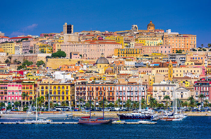 A deliciously inviting photo of a seaside town in Sardinia, shot from the ocean, showing a placid, blue water, sailing boats with sails furled in the foreground, beautifully coloured buildings rising up the hillside topped by a brilliant, bright, blue sky. 