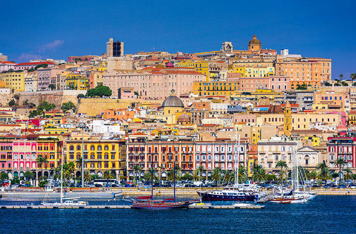 A deliciously inviting photo of a seaside town in Sardinia, shot from the ocean, showing a placid, blue water, sailing boats with sails furled in the foreground, beautifully coloured buildings rising up the hillside topped by a brilliant, bright, blue sky.