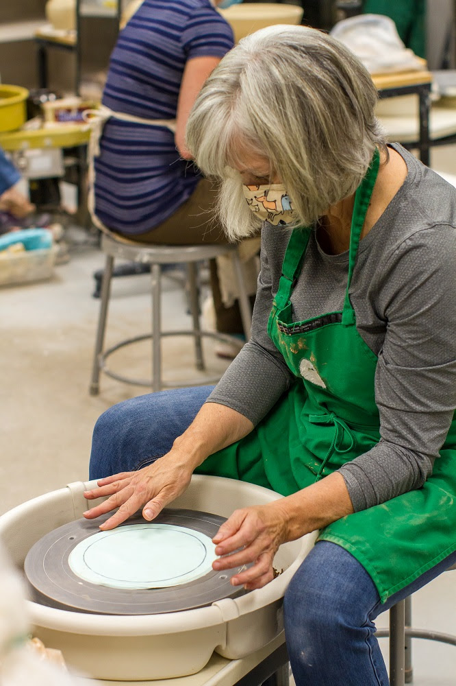 A white woman with grey hair sits at a pottery wheel
