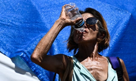 A very thin woman raises a plastic bottle to her lips, in full sun with a blue tarp behind her.
