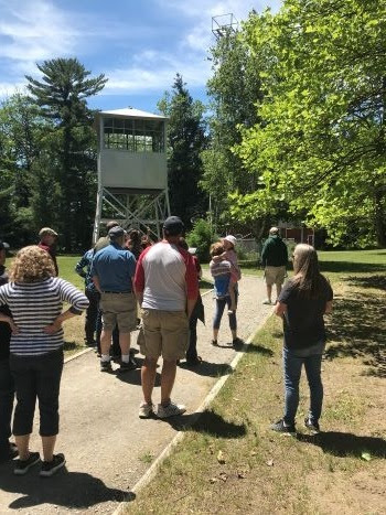 a group of men, women and children dressed summer attire stand on paved road, looking up at a tall, wooden fire tower in the forest