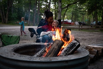 A woman with dark hair holds her face close to a smiling, blue-eyed little boy in her lap, in front of an orange campfire in a metal ring