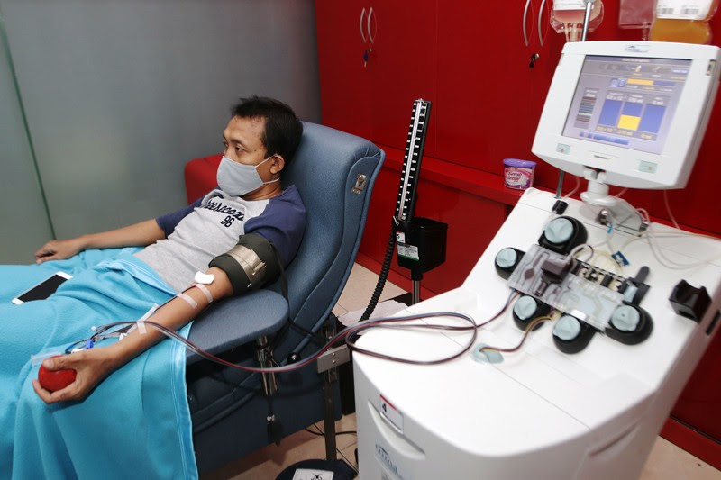A man wearing a face mask sits in a hospital bed while blood is drawn from his arm and plasma collected by apheresis apparatus.