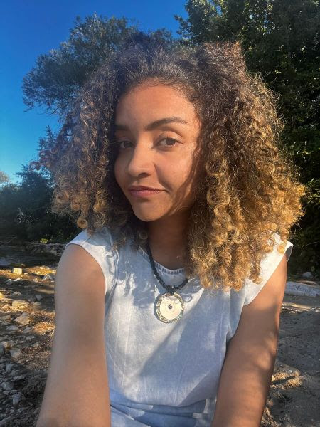 Larissa é ex-estudante do prouni. Ela tem cabelo cacheado com luzes loiras, veste camisa branca e colar.