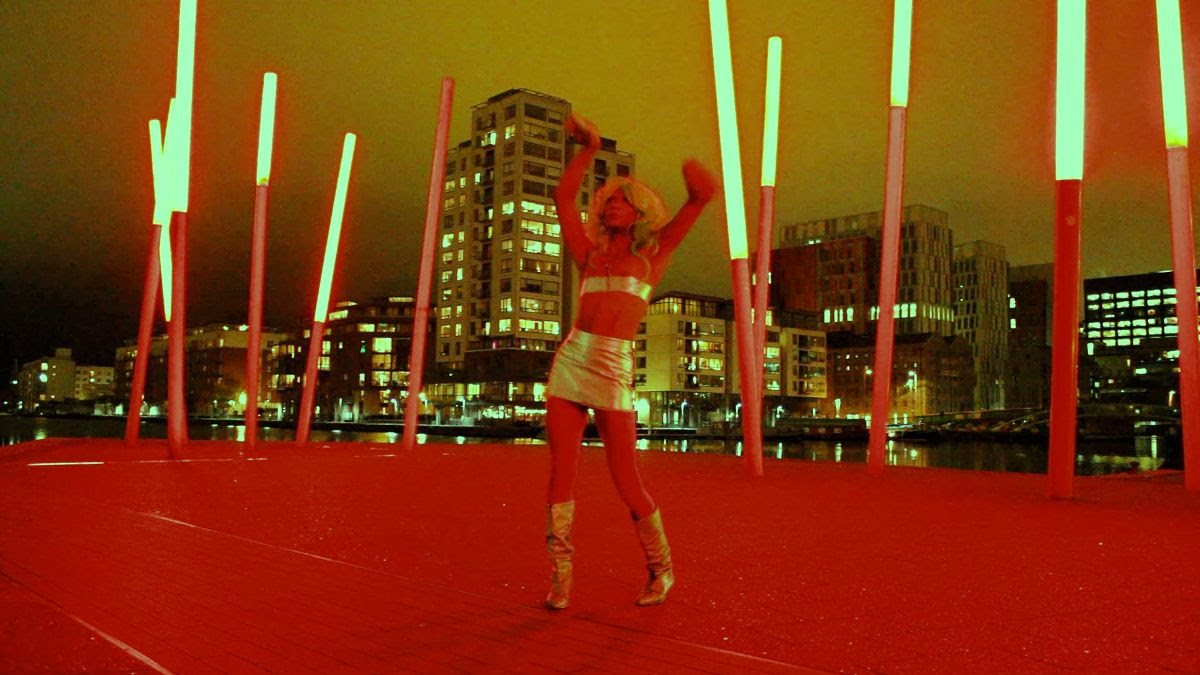 Venus Patel, wearing a gold two-piece mini skirt and belt top with gold boots, poses at the docklands in Dublin. The architectural lighting and city lights behind shine brightly the dramatic red-coloured ground that Venus is standing on reflects red colour onto their body