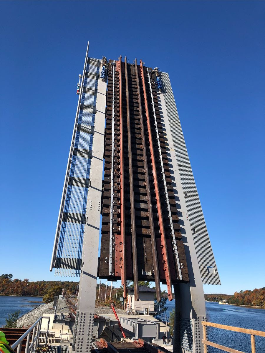 The movable bascule span of the drawbridge is shown at center and is vertical. Controls can be seen on the platform below. , perpendicular to the water below it. Commuter Rail tracks run down the center of the vertical span