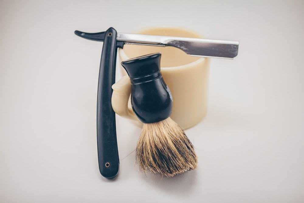 rasoio nero accanto a tazza in ceramica beige e pennello per crema da barba