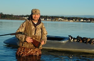 The Michigan DNR's Russ Mason in hunting gear, standing in the water holding a shotgun next to a boat with recently shot ducks.