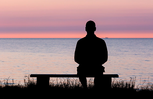 A silhouette of a man sitting on a bench looking out at the water. 