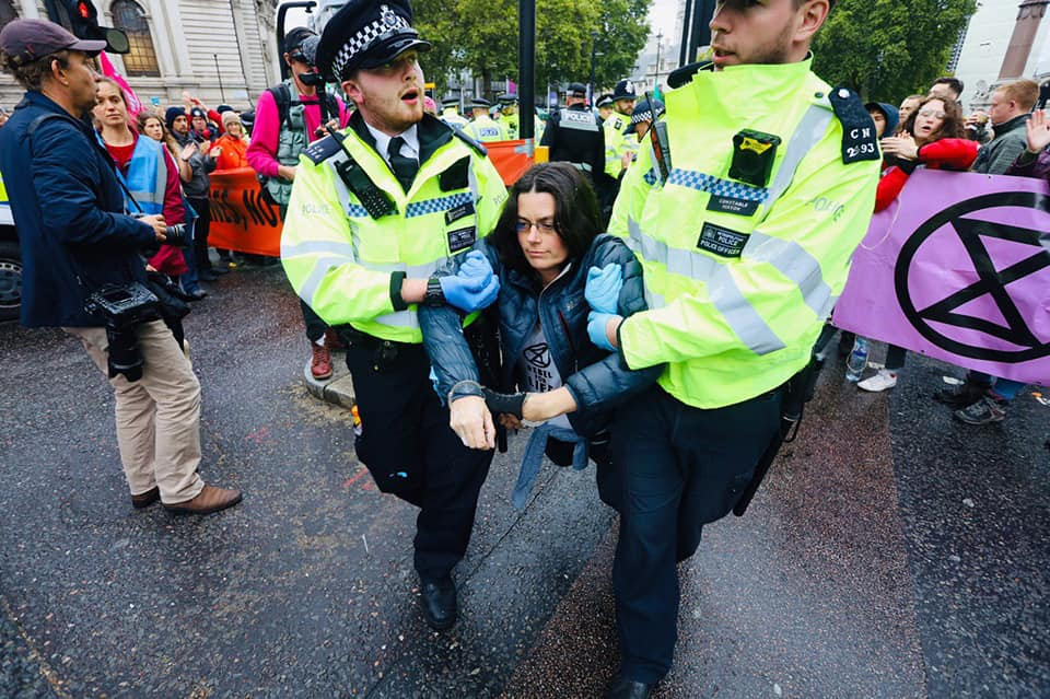 A woman being arrested and carried away by three police officers, she is wearing a t-shirt which says 'Rebel for Life' on it.