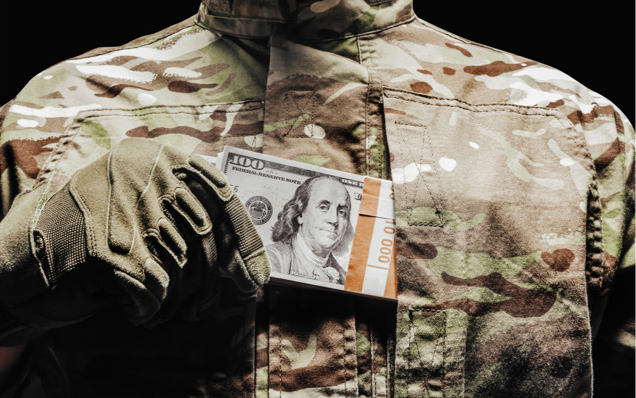 A close up on the chest of a person wearing camouflage gear, surreptitiously sliding a bundled stack of $100 bills into a pocket