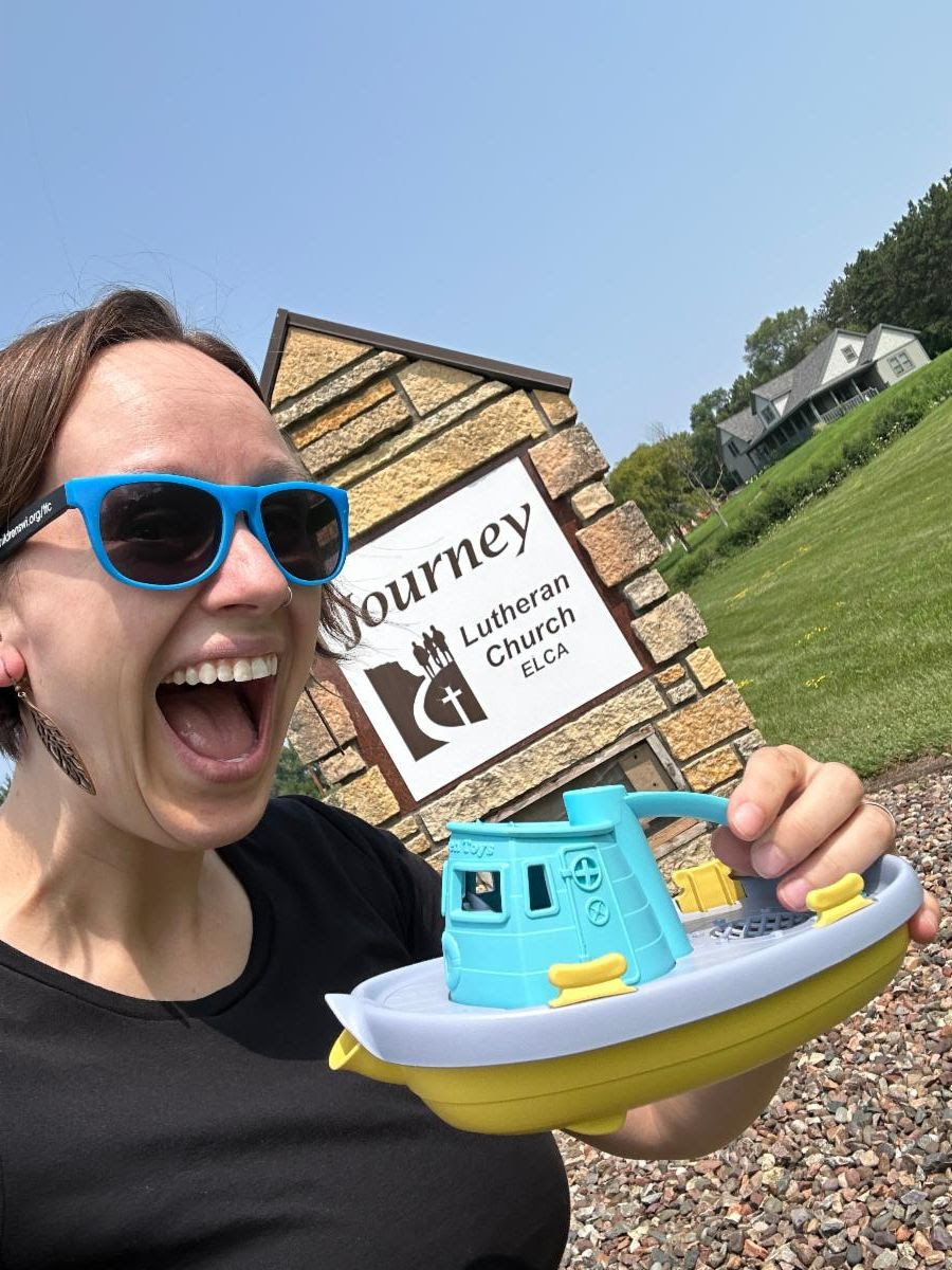 A gleeful picture of Breanna holding a toy "ecumenical" boat, while standing in front of the sign for Journey Lutheran Church, on a sunny day