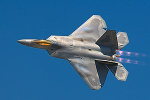 F-22 Raptor banking left in-flight, showing the top view of the aircraft. The engines with afterburners emit a pinkish glow. Aircraft mostly gray, apart from the gold cockpit window, with hints of bluish condensation on the wings.