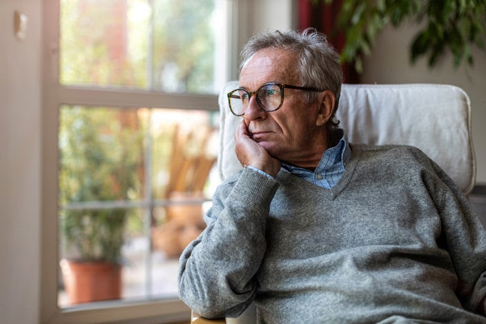 Older man in glasses and a sweater sitting in a living room chair looking out the window.