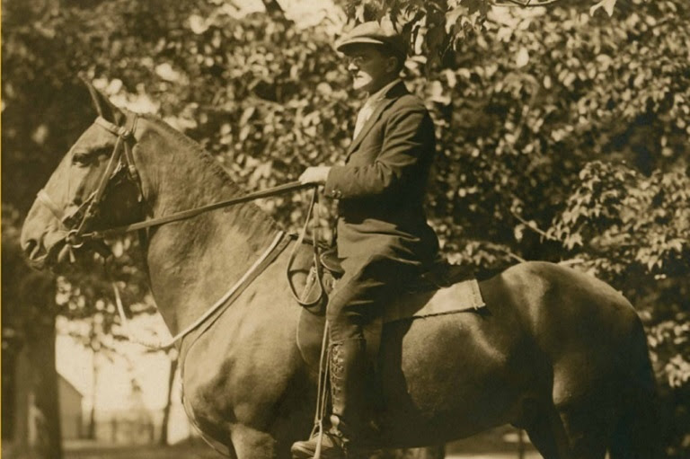 Sepia photo of a man wearing a paper boy hat riding a horse.