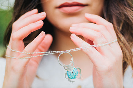 A photograph of a model holding a silver necklace by Ailsa Ritchie. The model is wearing white and only their hands and chin are visible. The necklace has a long silver chain and a pendant that is a silver beetle with blue details. 