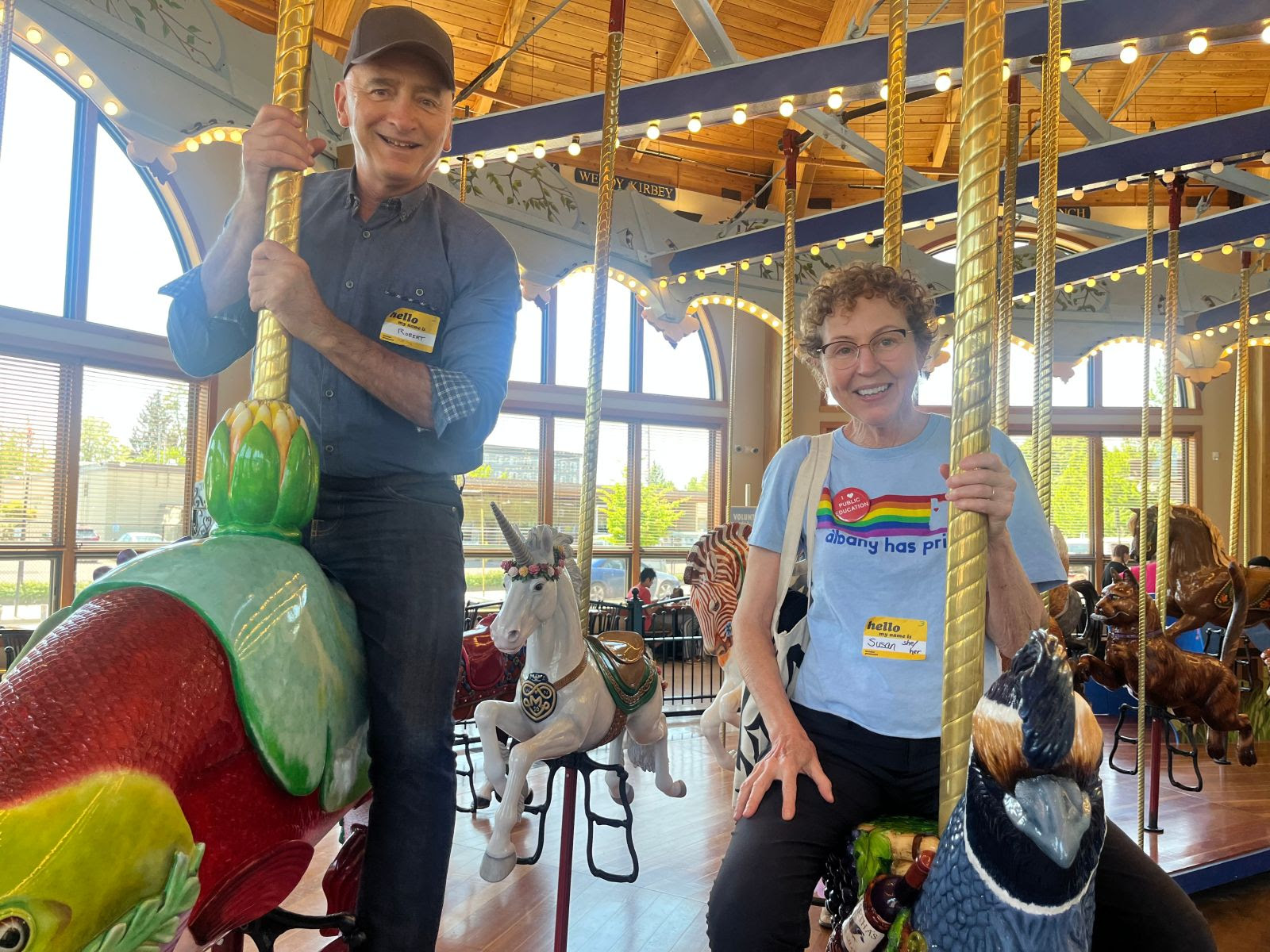 Robert Bray and Susan ride the Albany carousel with bright windows behind them.