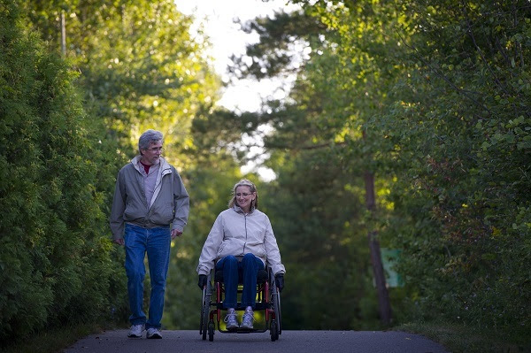 an older couple, the man wearing jeans and zip-up jacket and the woman in jeans and a light sweater, also in a wheelchair, on a paved path
