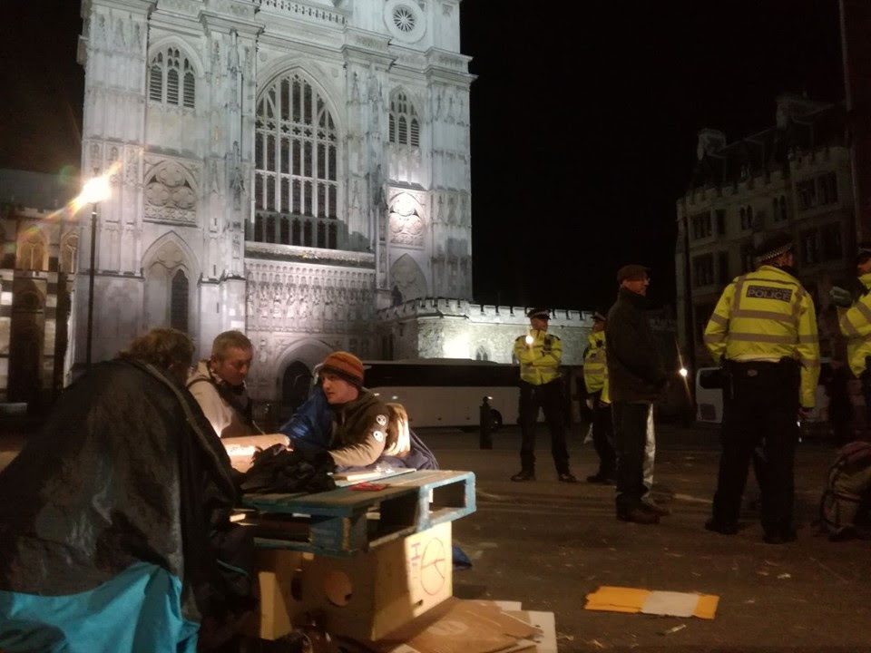 3 rebels, including Callum, sat on the ground in front of Westminster Abbey. Two of them have their arms locked into a tube. It is dark, and there are 4 police officers stood around them.