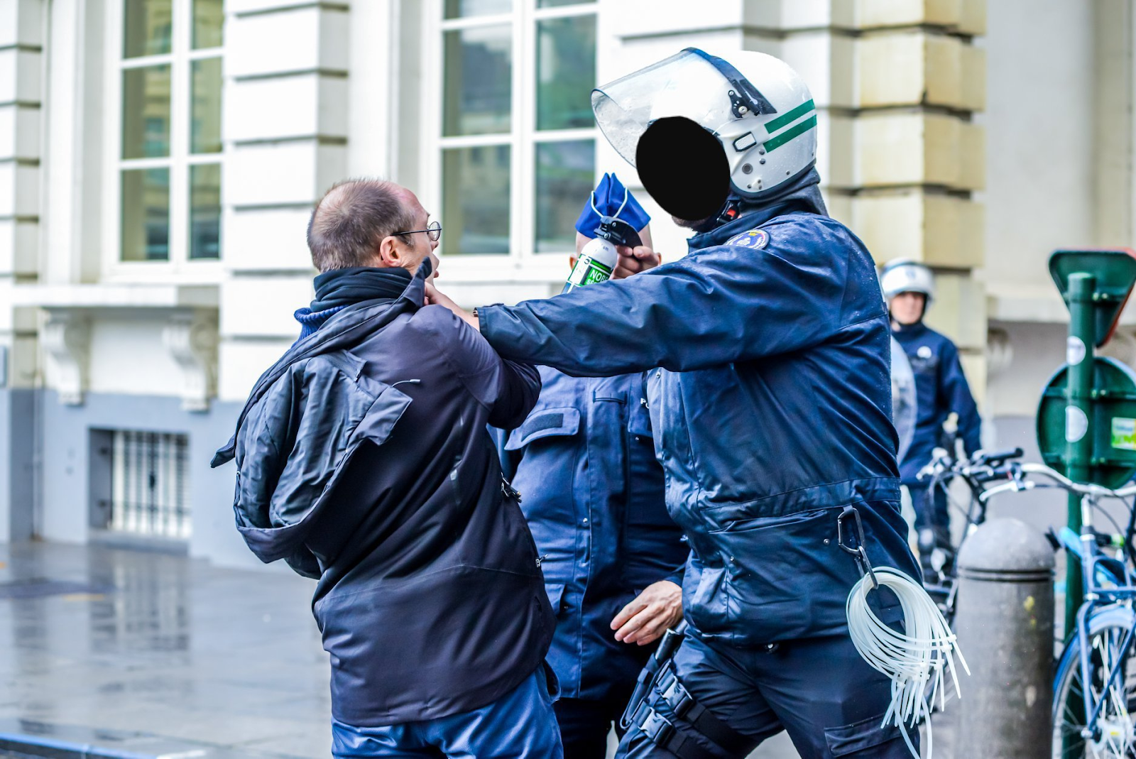 A rebel being held by the neck by a police officer. In his other hand, the police officer has a bottle of pepper spray which he is pointing at the activist.