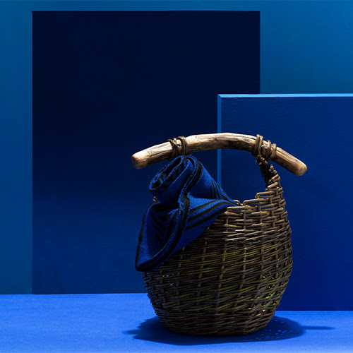 A photograph of a willow basket by Naturally Usefulagainst a dark blue background. There is a dark blue and black wool scarf by Green Thomas in the basket.