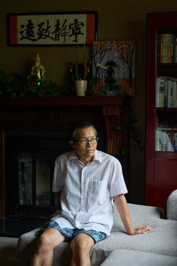 Deng Yuwen wears a short-sleeved button down shirt and sits on a gray sofa while looking into the distance.