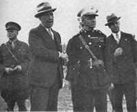Five men, two in the foreground and three in the background, one mostly obscured. Two men are in suits and three are in their military dress uniforms. All of the men in the picture are wearing hats. The two men in the foreground are shaking hands.