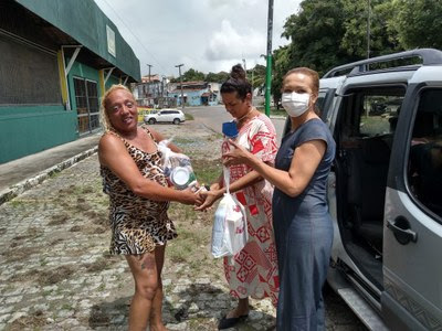 A imagem mostra três travestis,uma delas usando máscara de proteção entregando um pacote com alimentos.