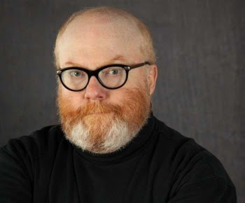 An older man with a ginger and white beard, thick glasses and inviting expression.