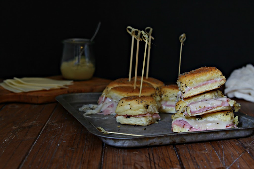 Hot Ham and Cheese Sliders on baking tray with decorative toothpicks. Small glass jar of mustard with spoon and slices of cheese on cutting board in background.
