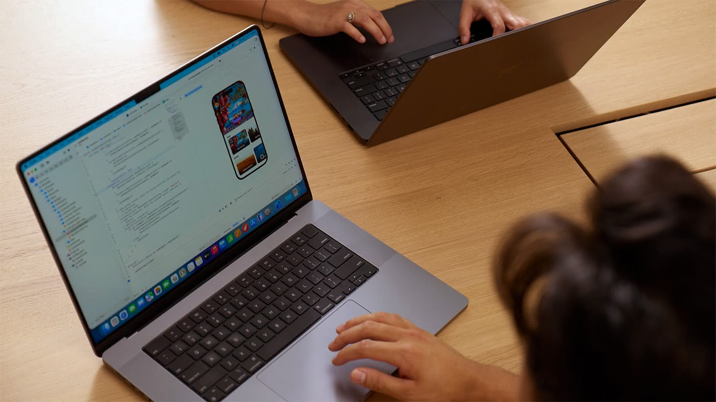 An overhead view of two people working side by side at a light wood table, each on a MacBook Pro. The foreground laptop displays Xcode with an iOS app simulator showing a colorful travel or photo-browsing app on a virtual iPhone screen. The second person's hands are visible in the upper portion of the frame, typing on their MacBook, suggesting a collaborative development or code review session.