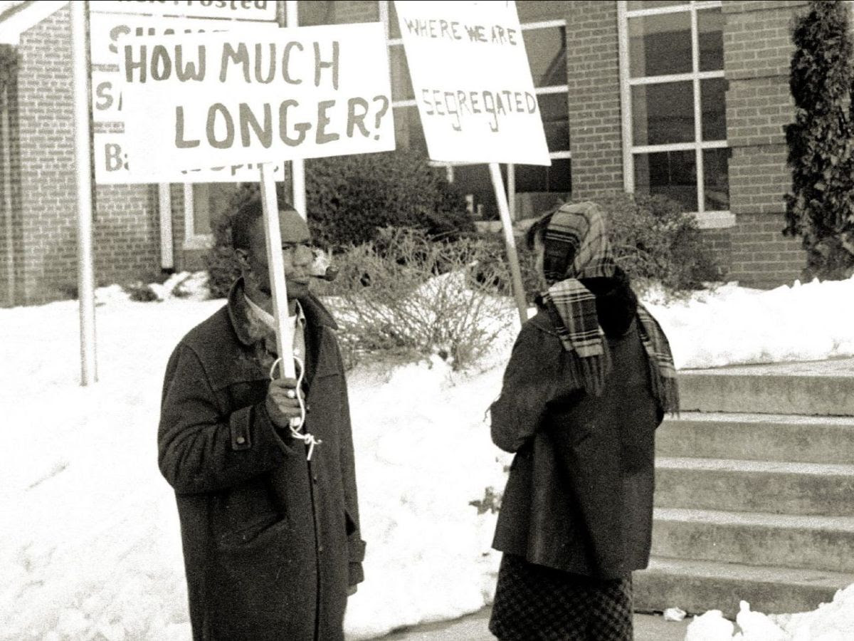 A black and white photograph shows two Black people in front of a brick building. The people are holding signs. One sign reads "How much longer?" The other sign has some visible words: "Where we are segregated." The people are wearing coats and scarves. There is snow on the ground.