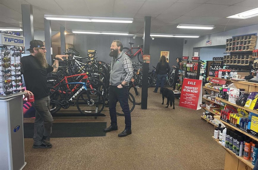 Two people stand and talk in a bicycle store. Bikes are standing up on display, along with sunglasses, bug spray and sunscreen.