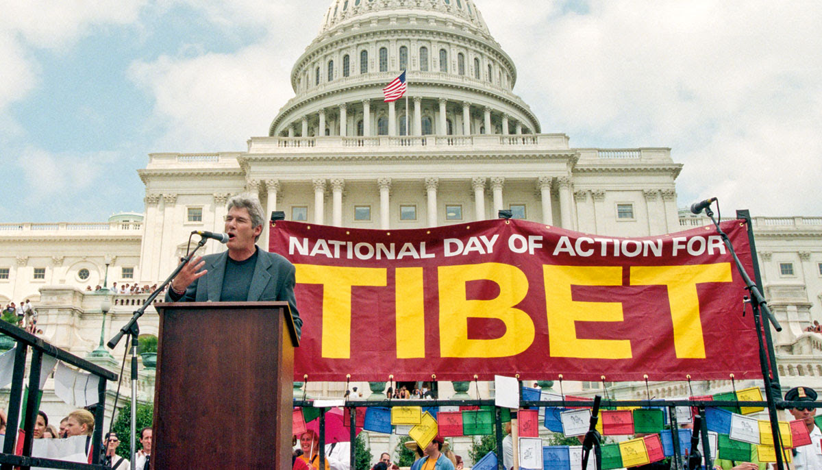 A man stands at a microphone with a sign in the background that reads "National day of action for Tibet."