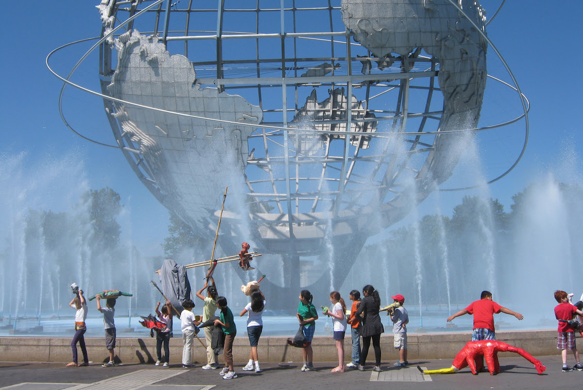 A group of children are standing in line in front of the Unisphere holding sculptures they have made at the Queens Museum. Water jets are surrounding the Unisphere and are shooting upwards. 
