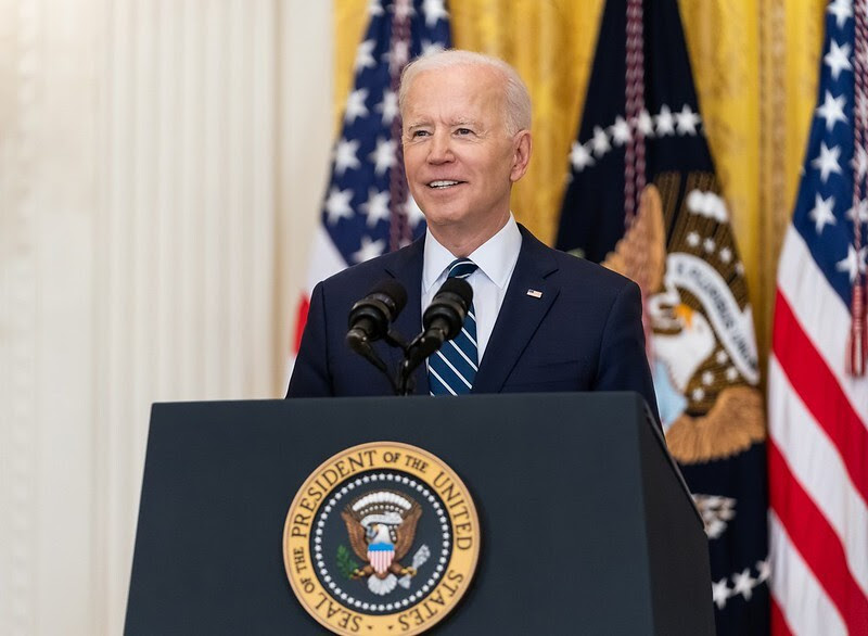 Joe Biden é um homem branco na faixa dos 70 anos, ele tem cabelos grisalhos; na foto Biden veste terno e gravata azul e sorri enquanto posa em cima de um palanque