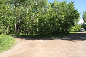 view of a partially forested area, with a wide gravel clearing, and a camper parked in the back on the right