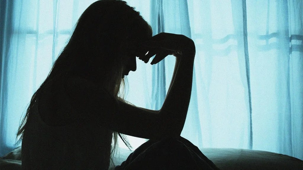 there is the silhouette of a person sat against a blue background of white curtain, with their head resting on their hands