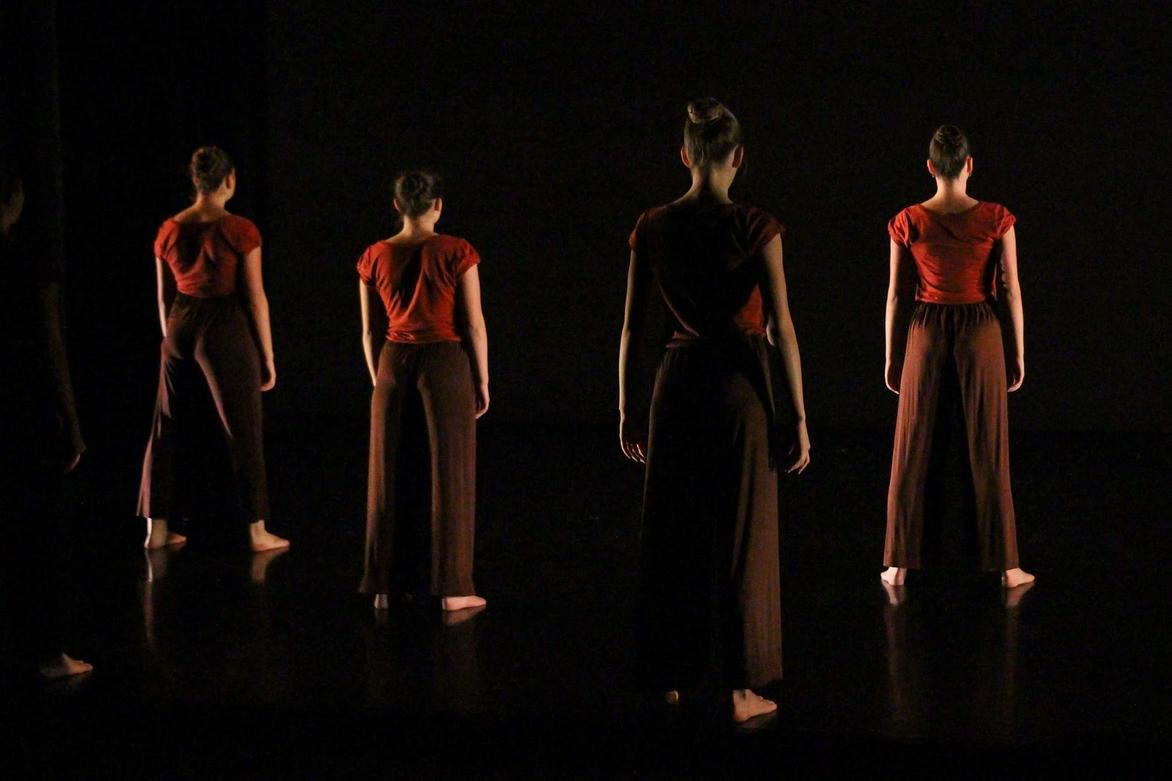 Four dancers stand with backs to the camera in dramatic darkness