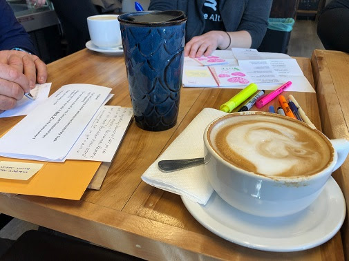 Two people sit at a table with their coffee, writing postcards.