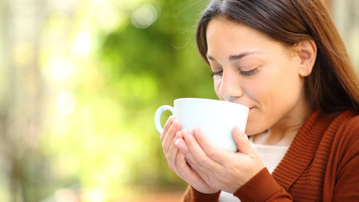 Close-up on a woman drinking a cup of tea. She is smelling and looking down at the white cup with a lush green background behind her.