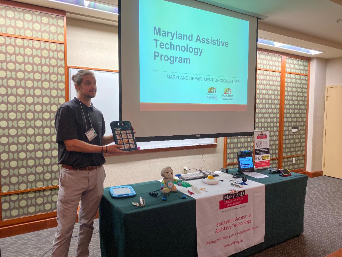 AT Program employee, a young white man with brown hair pulled back in a black shirt and slacks displays an AAC device. He stands beside a projection screen reading "Maryland Assistive Technology Program - Maryland Department of Disabilities" below which is a table full of various Assistive Technology items