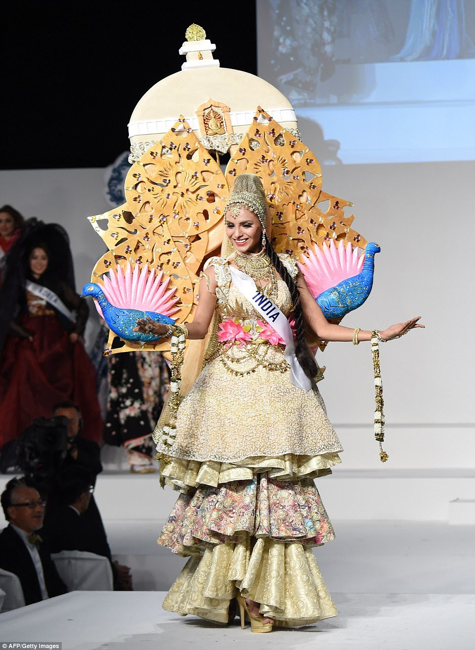 Miss India Supriya Aiman wore a heavy-looking pagoda-inspired outfit based on the Vishwa Shanti Stupa in Delhi. Her outfit also featured peacocks, India's national bird, although this time they had pink spiky feathers like a lotus flower. The gold tear-drop shapes were decorated with cut-out hearts and lead up to the main dome, which was decorated with a Buddha statue. Supriya also wore an intricate multi-layered gold dress with a water-lily and gold chain embroidered waist belt, a heavy gold collar necklace and embroidered headdress